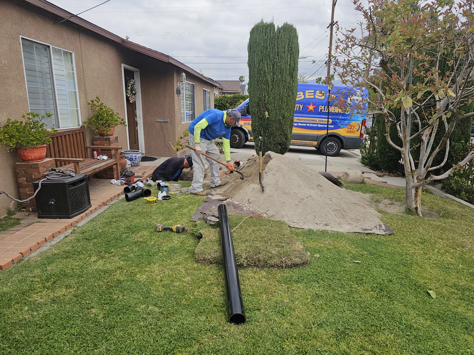 Plumber at a residential excavation with replacement pipe staged on the lawn